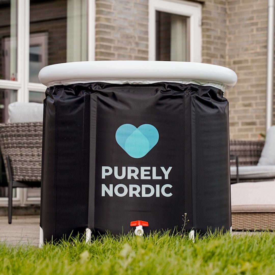 En sort, bærbar Isbad Deluxe 2.0 spa med "Purely Nordic" og et blåt hjertelogo på siden, placeret på en græsplæne på baggrund af et hus med terrasse og udendørsmøbler.