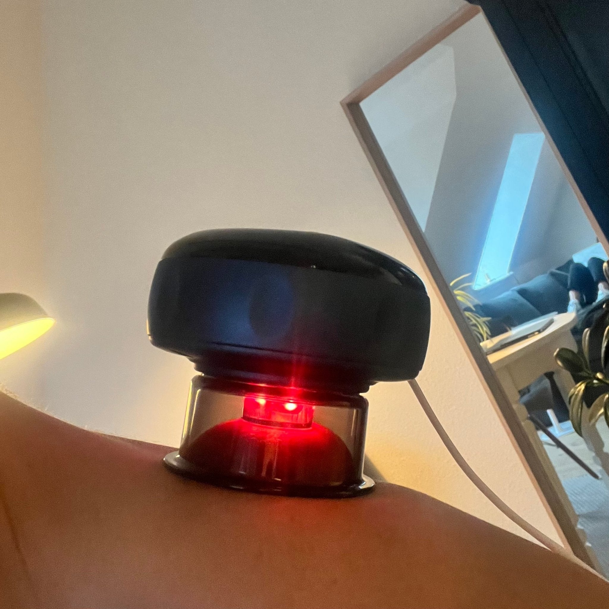 En Smart Cup Basic elektronisk massageanordning påføres en persons skulder. Enheden er sort og cirkulær og udsender et rødt lys. Den er placeret i et rent nordisk hjemmemiljø med spejl og del.